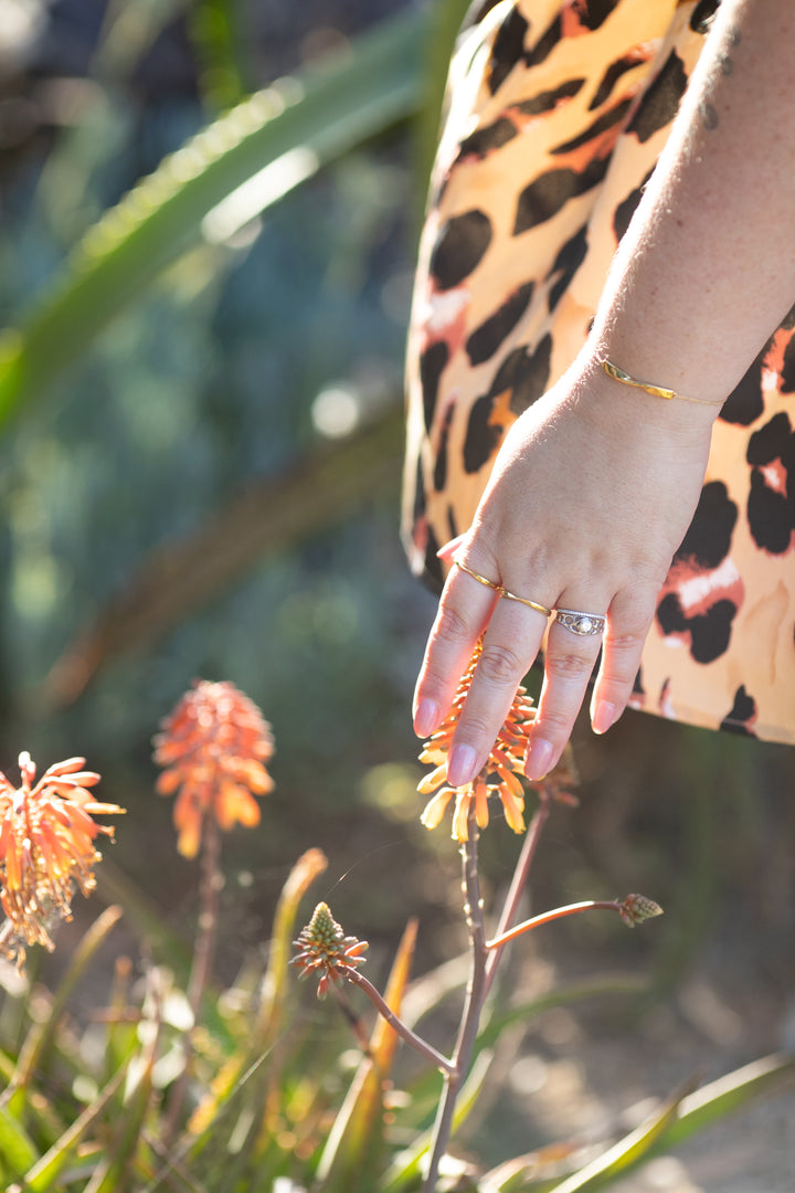 an image of a plus size woman's hand hovering over a flower with a twist ring in gold on both her pointer and middle fingers, and a twist bracelet in yellow gold on her wrist. Her leopard print dress billows in the wind behind her. #color_gold