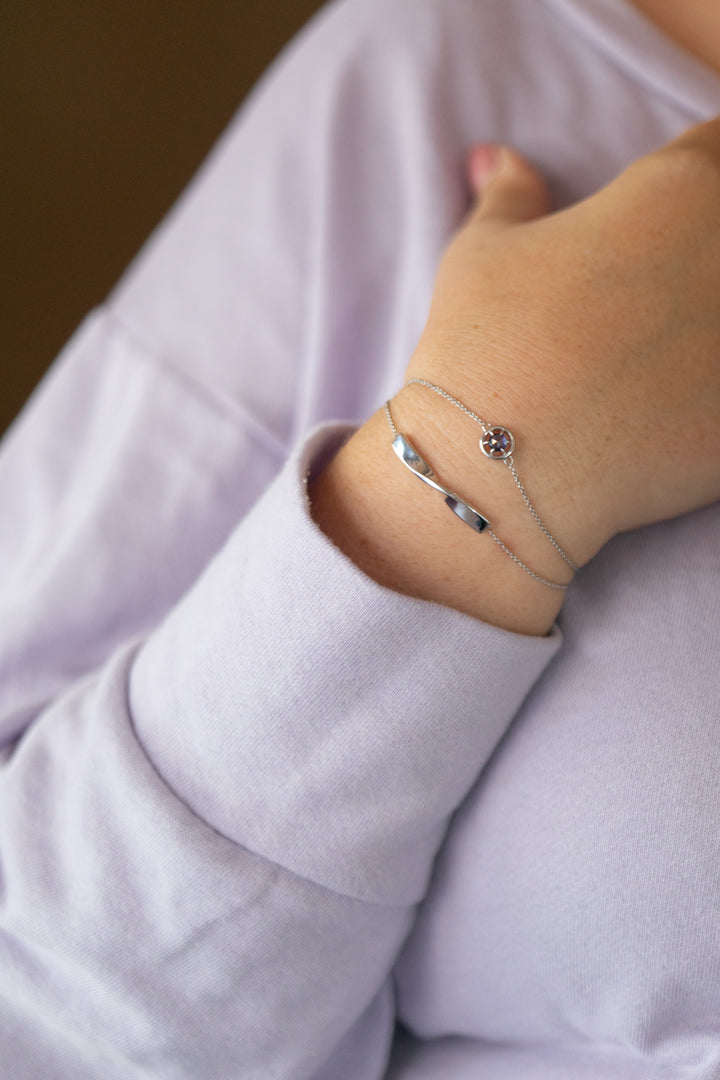 A stylized image of the gemstone bracelet in silver and tanzanite on a plus size woman's wrist and styled with the twist bracelet in silver. She is wearing a lavender colored sweatshirt.