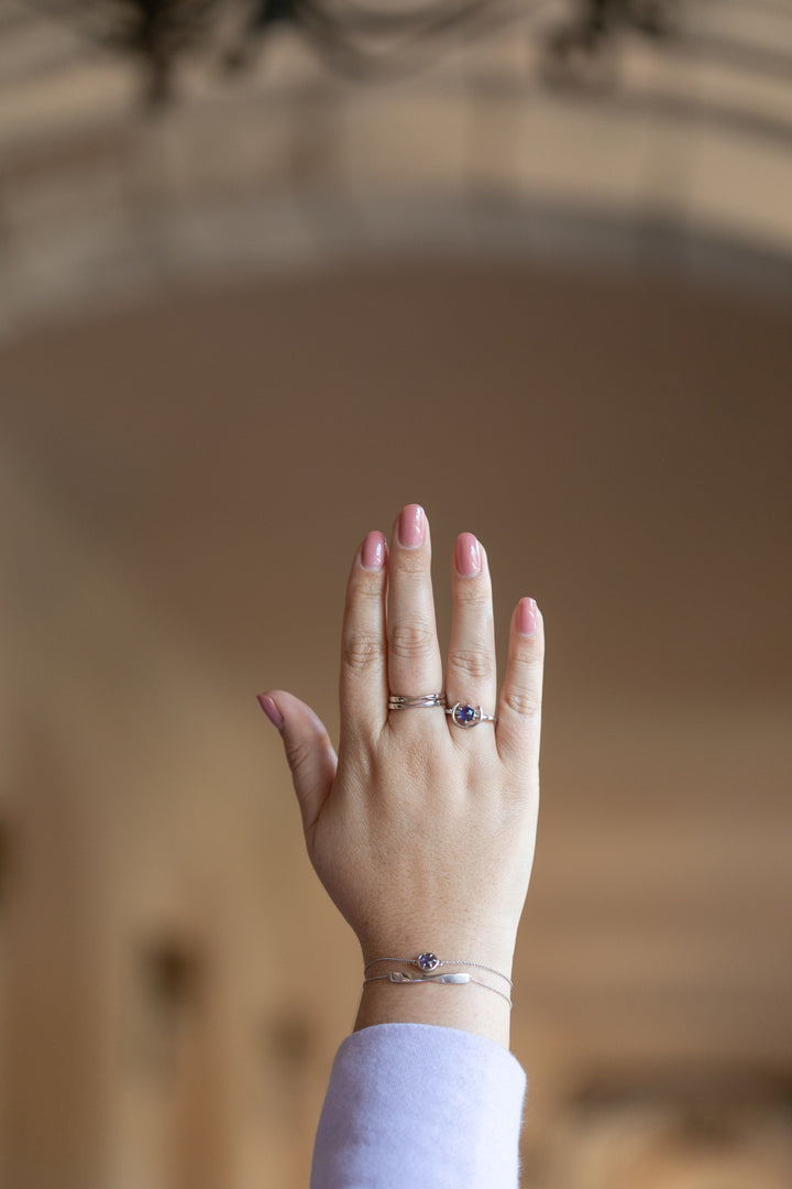 An image of a plus size woman's hand held up in a hallway wearing two silver twist rings stacked on her middle finger, and a gemstone ring in silver and tanzanite on her ring finger. She is also wearing the gemstone bracelet in silver and tanzanite with the twist bracelet in silver. She has light pink nail polish and a lavender sweatshirt on.