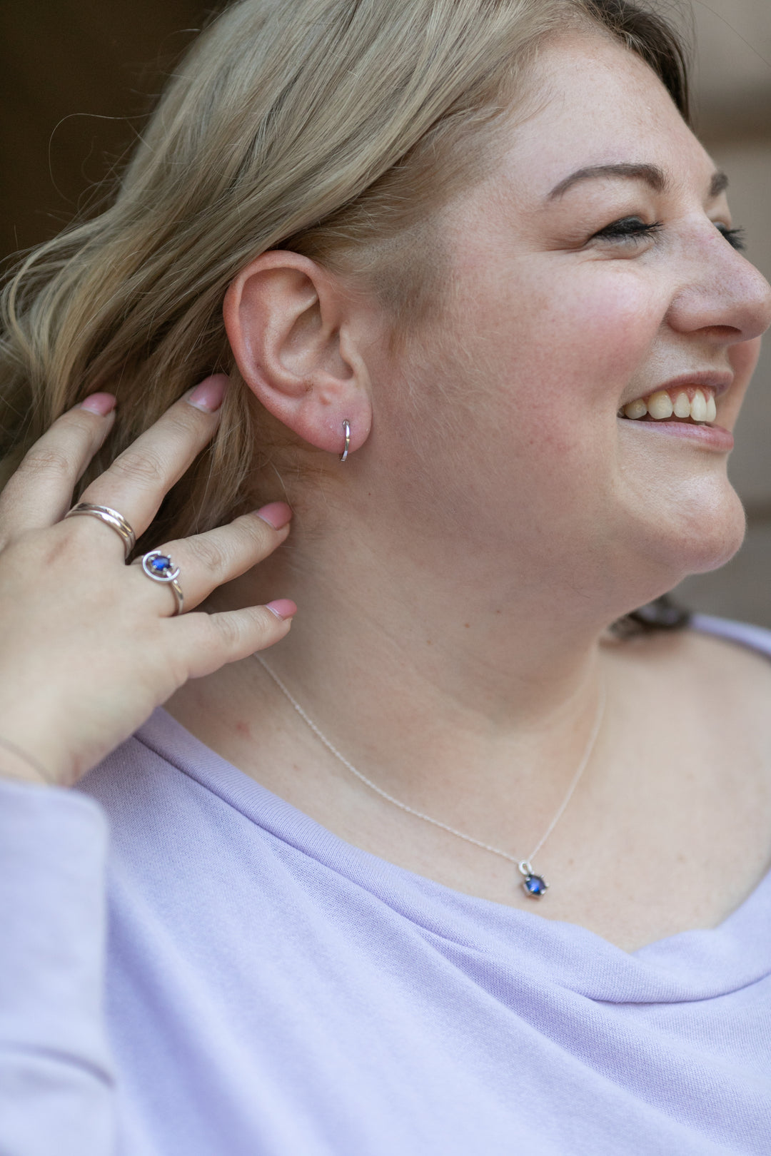 a smiling woman pulls her blonde hair back to reveal a simple huggie hoop earring in sterling silver on her ear. #color_silver