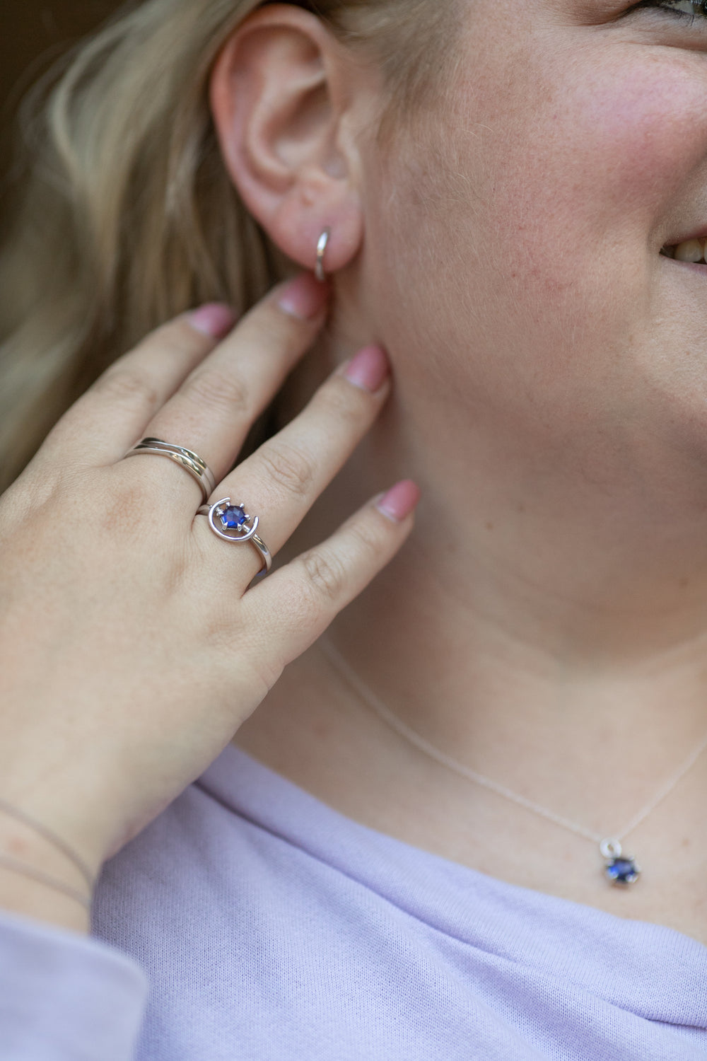 a close up shot of a blonde woman pushing her hair back to show off her simple huggie hoop earring in sterling silver. Her hands feature two twist rings on her middle finger and a gemstone ring in silver and tanzanite on her ring finger. #color_silver