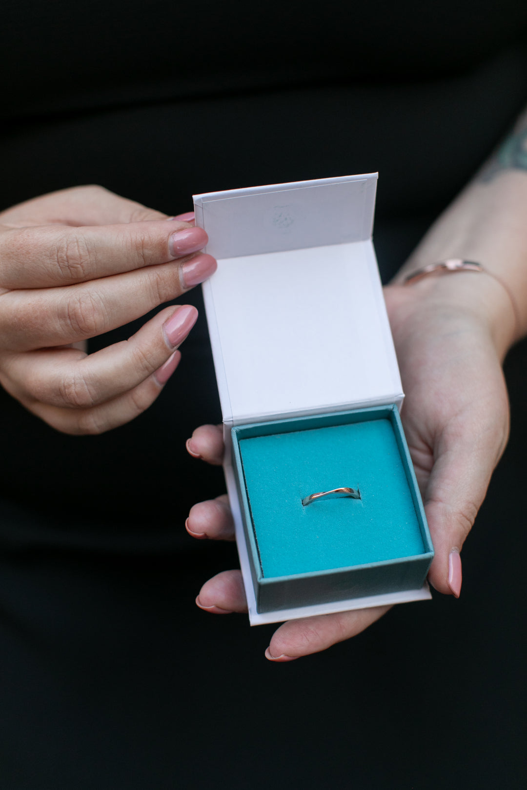 two hands hold open a Chouette Designs branded white and teal ring box to reveal a rose gold twist ring 
