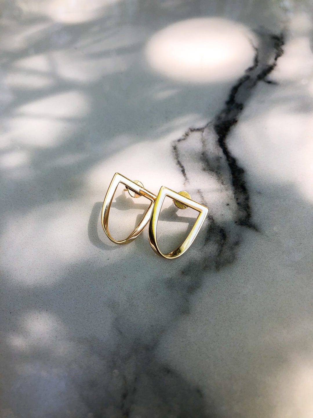 a pair of twist earrings in yellow gold sit atop a marble table top with speckled shadows across it. #color_gold