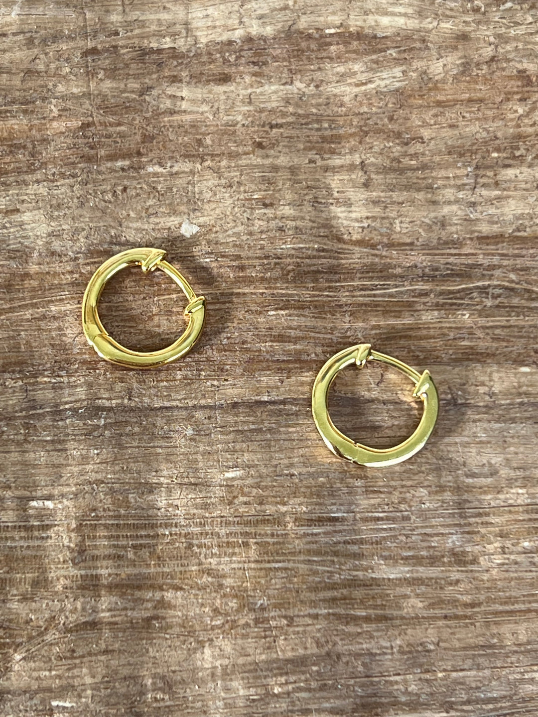a detail image of two huggie hoop earrings in yellow gold laying on top of a wood grain backdrop. #color_gold