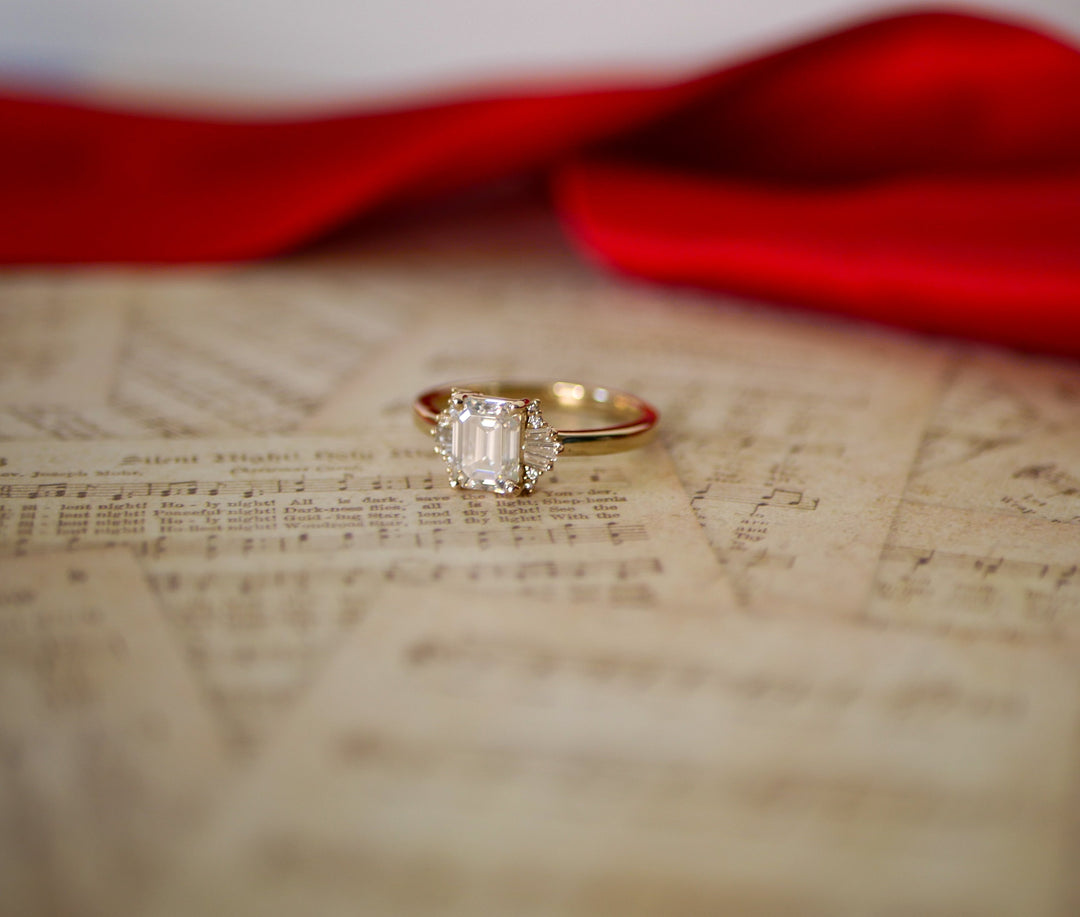 a gold ring featuring an Emerald cut moissanite, as well as round and tapered baguette cut diamonds sit atop sheet music. A red ribbon is visible in the background