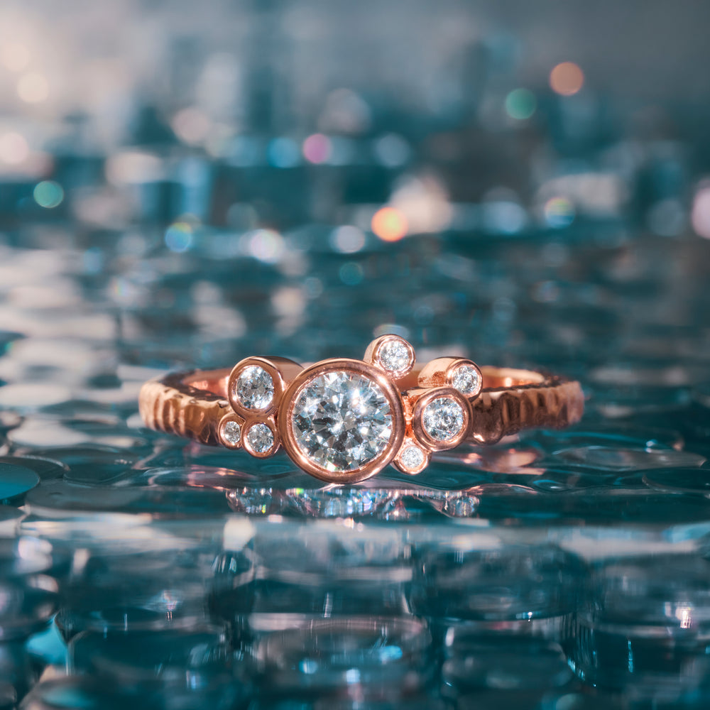 a rose gold engagement ring with bezel set round diamonds appears to be floating on water
