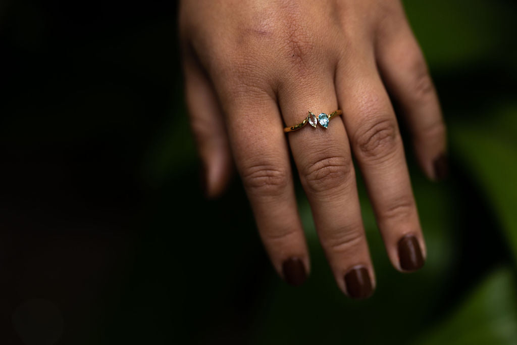 A close up image of a hand wearing the Monteverde Ring- a moi et toi style ring with a marquise cut green amethyst and a pear cut blue zircon set in a gold hammered band.