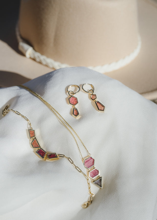 the earrings styled in a flat lay alongside the warm ember bracelet and the 3 stone pendant on a white piece of fabric. A neutral felt hat is in the background