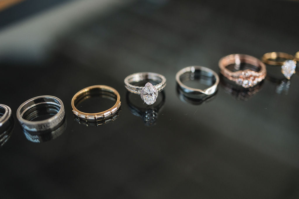 A row of various styles of rings in a row atop a black background. The central ring in focus is a white gold oval cut solitaire style