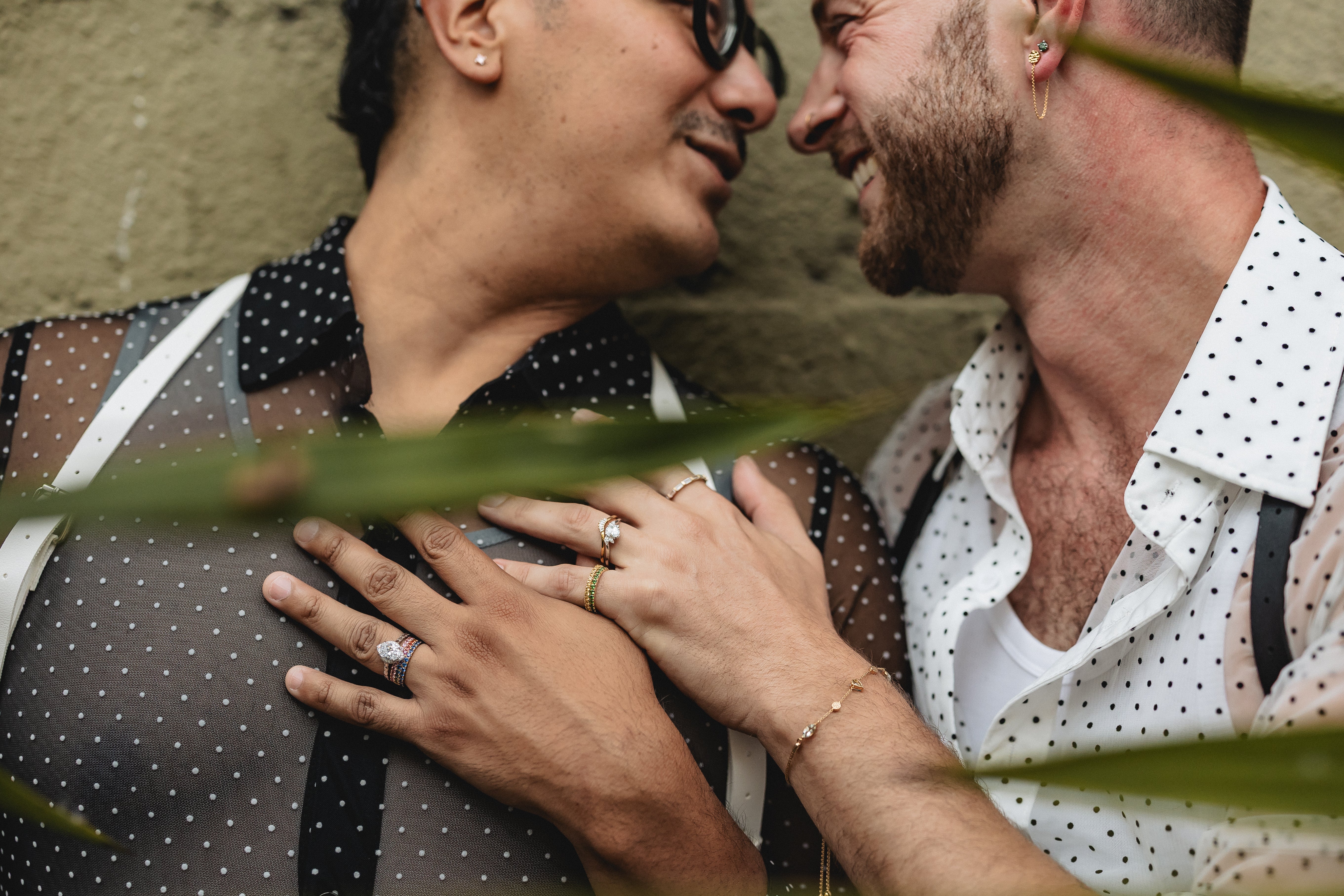 Two men embracing with a close-up of their hands and faces, surrounded by green leaves.