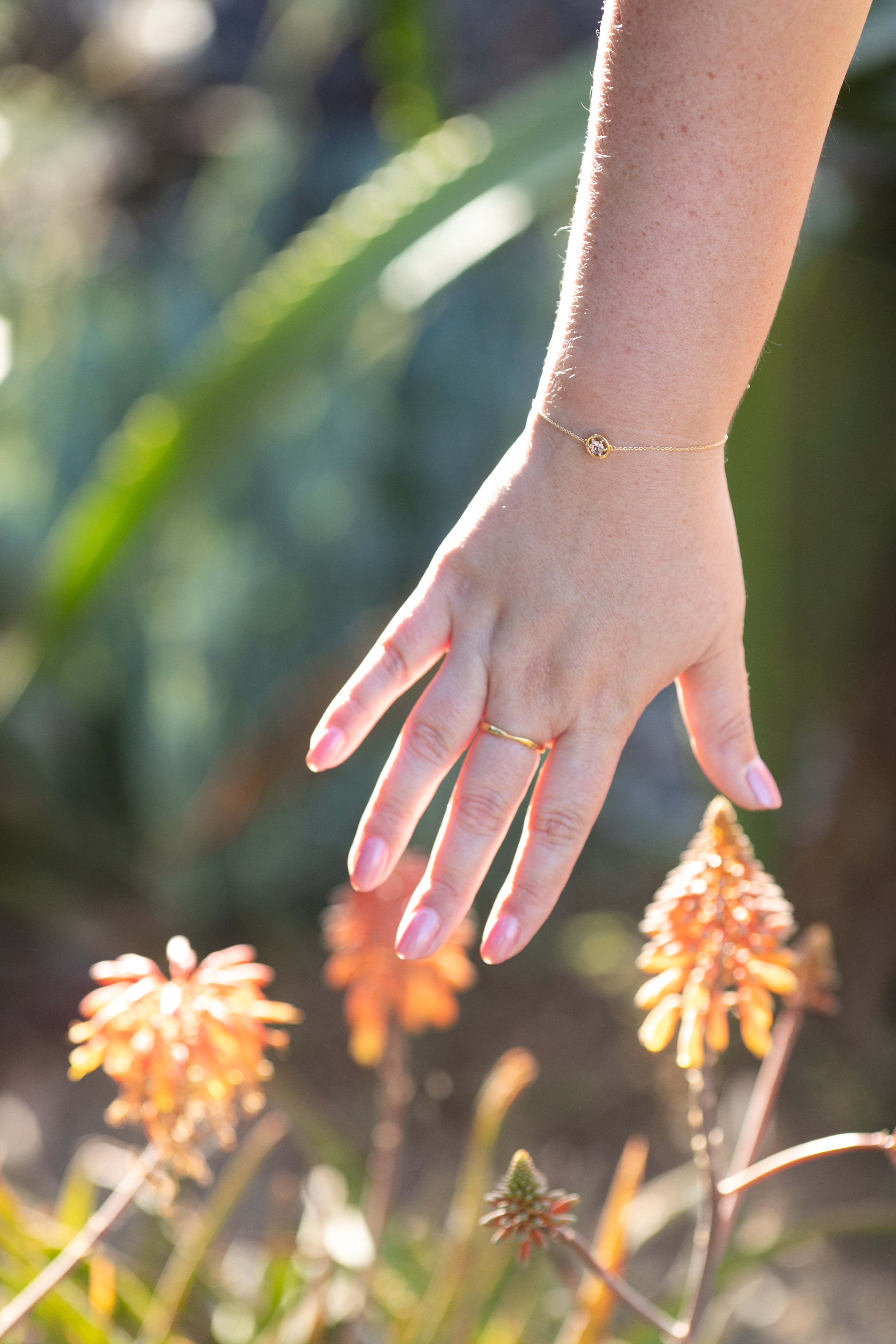 A shot of a plus size woman's hand in front of orange flowers wearing the gemstone bracelet in gold and spinel styled with a simple gold twist ring on her middle finger.