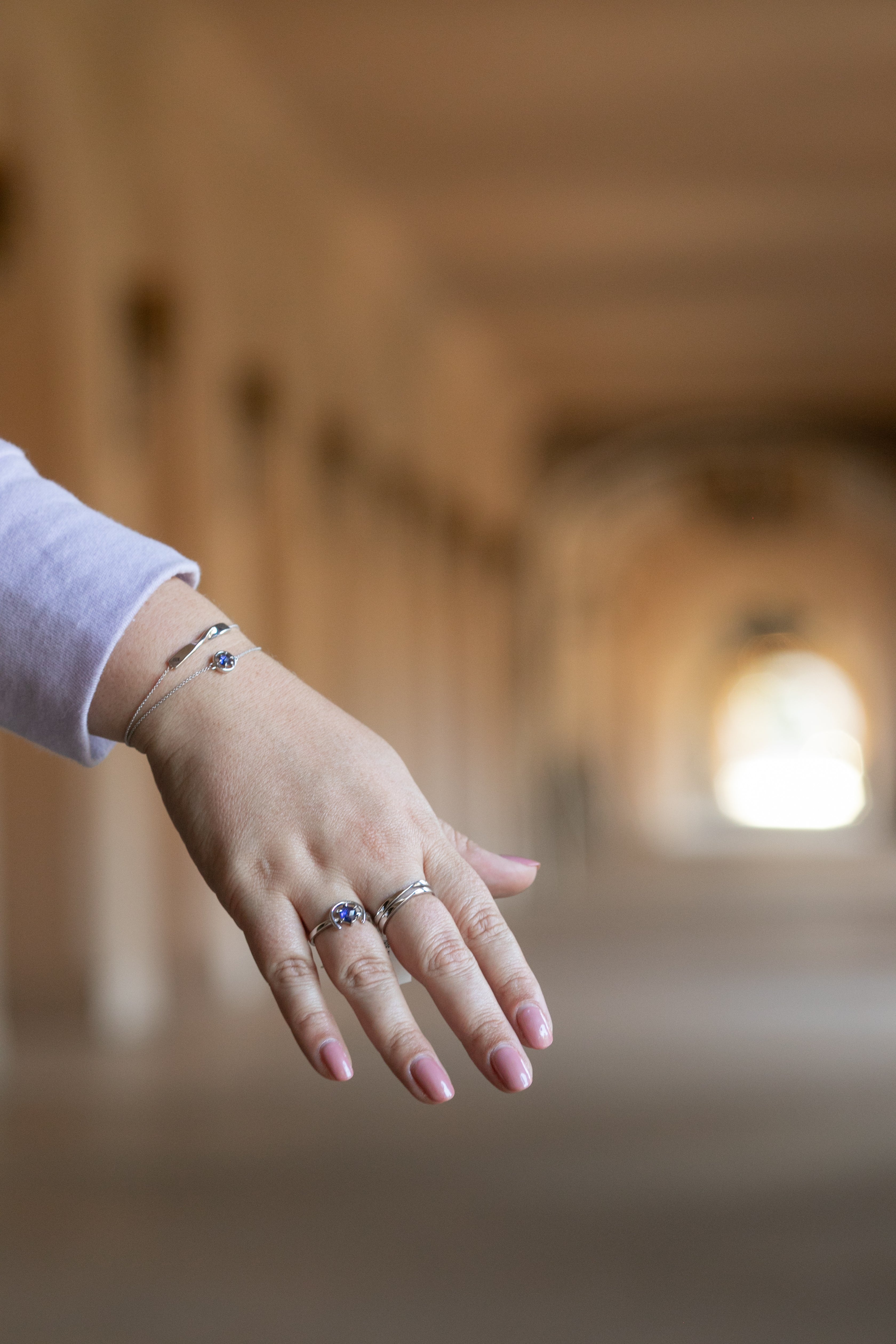 A hand with a hallway in the background. There are two silver stacked twist rings on the middle finger, a silver and tanzanite gemstone ring on the ring finger and two silver bracelets on the wrist. Plus size and size inclusive rings and bracelets.