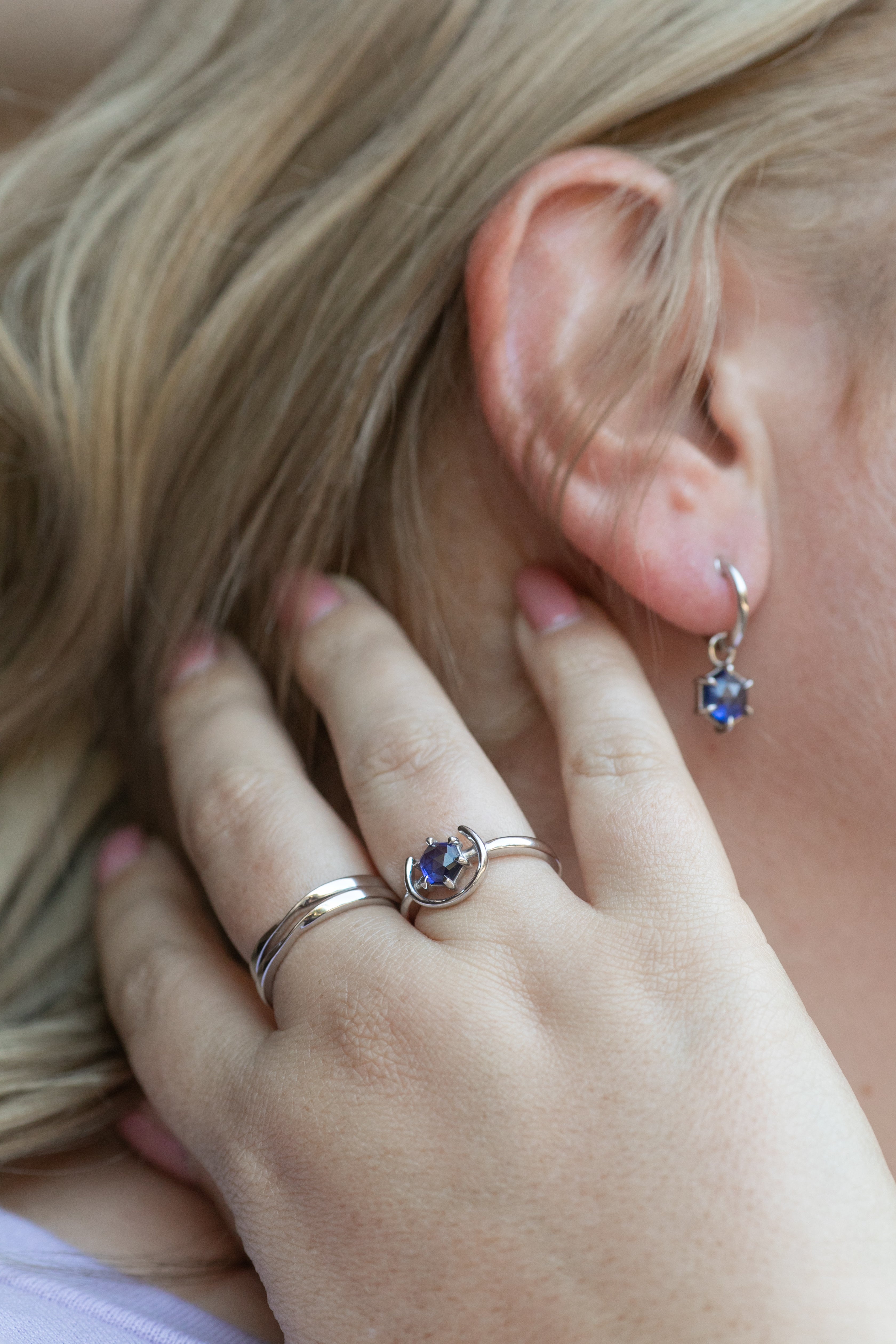 A close up shot of a blonde woman's ear wearing a silver and tanzanite gemstone huggie hoop earring. Her hand is rested near her hair, wearing two silver twist rings stacked on her middle finger and a gemstone ring in silver and tanzanite on her ring finger.