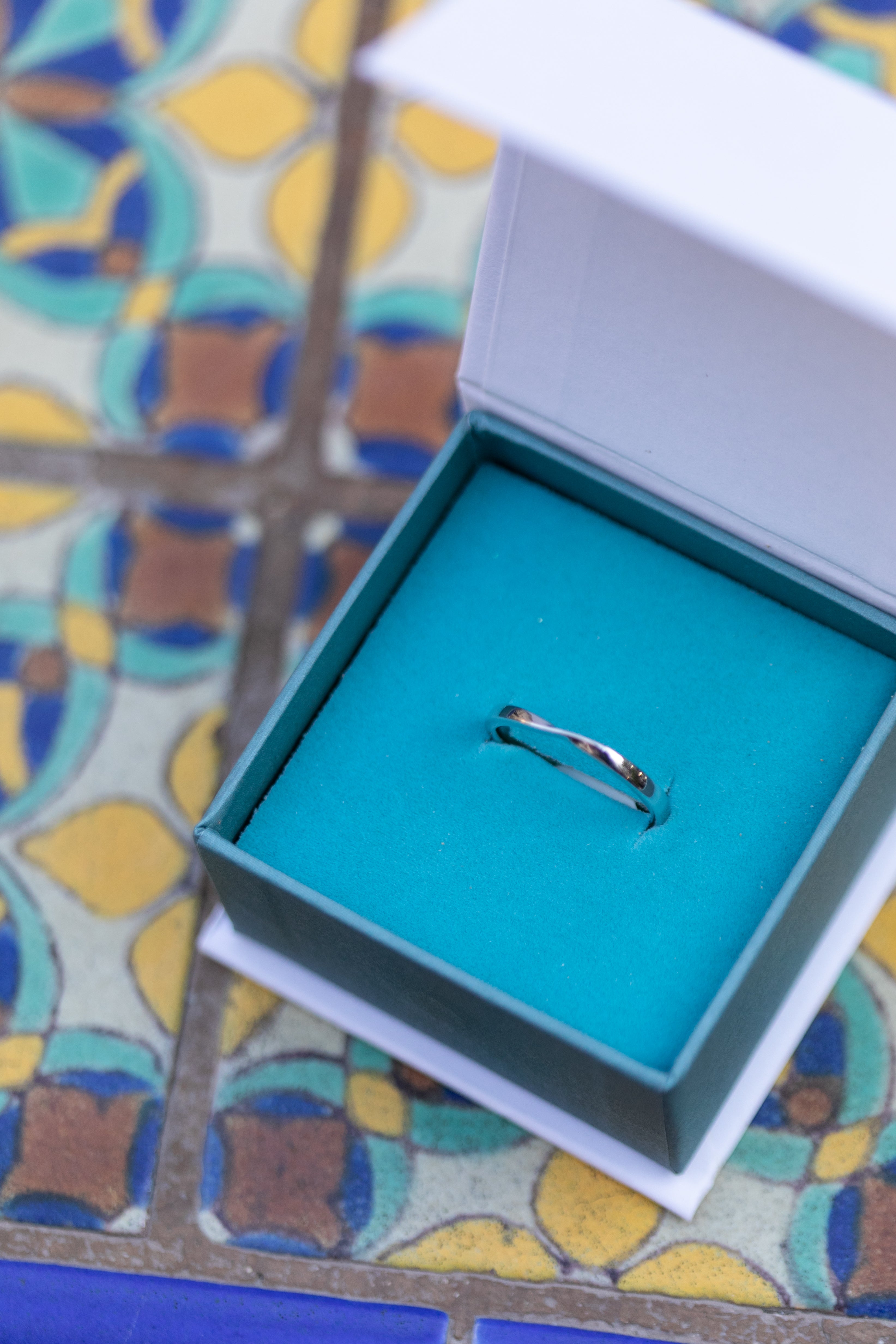 A close up of a silver twist ring in a Chouette Designs branded teal and white ring box, sitting on top of colorful Spanish style tile.