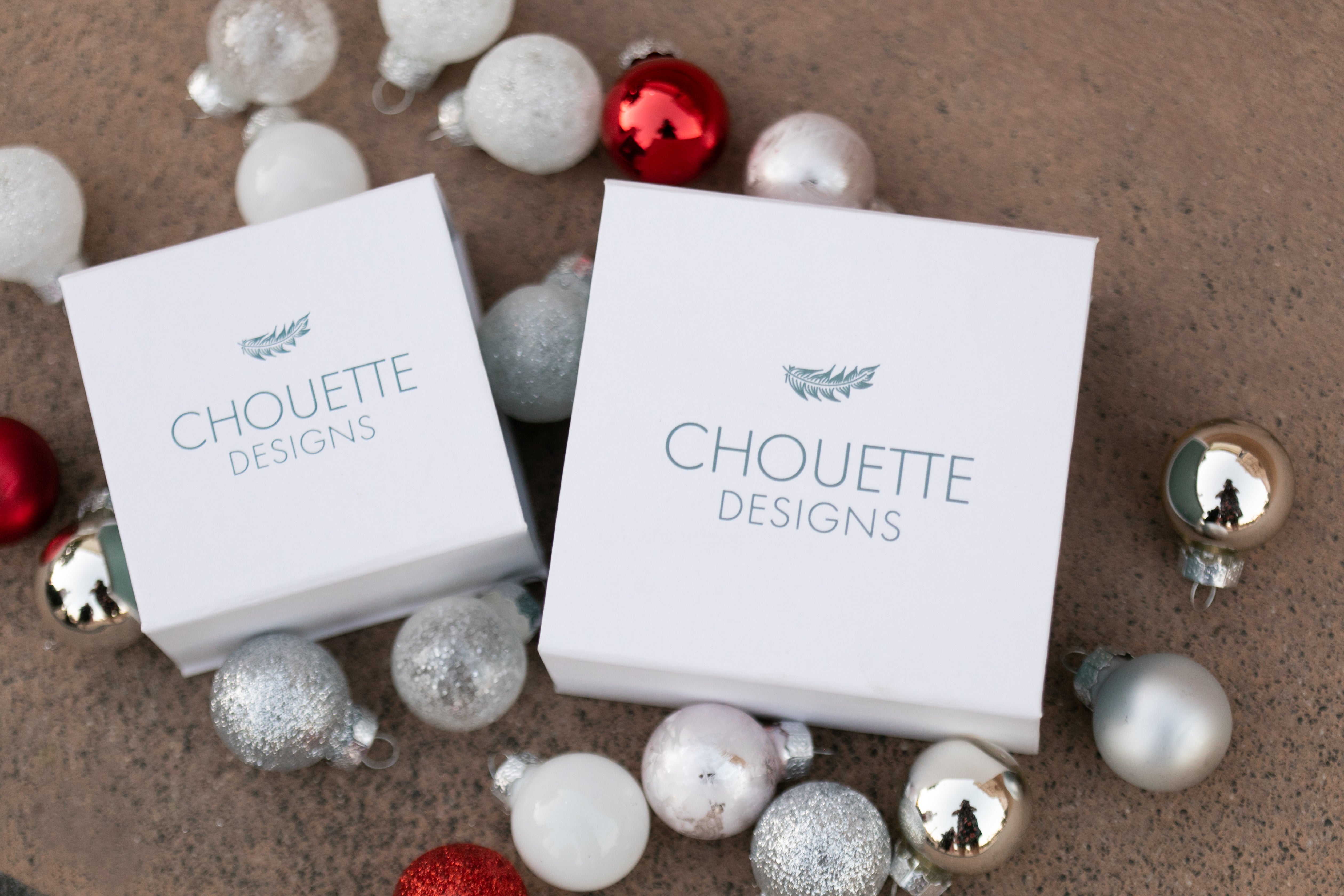 Two Chouette Designs branded jewelry boxes placed among red, white and silver Christmas ornaments.