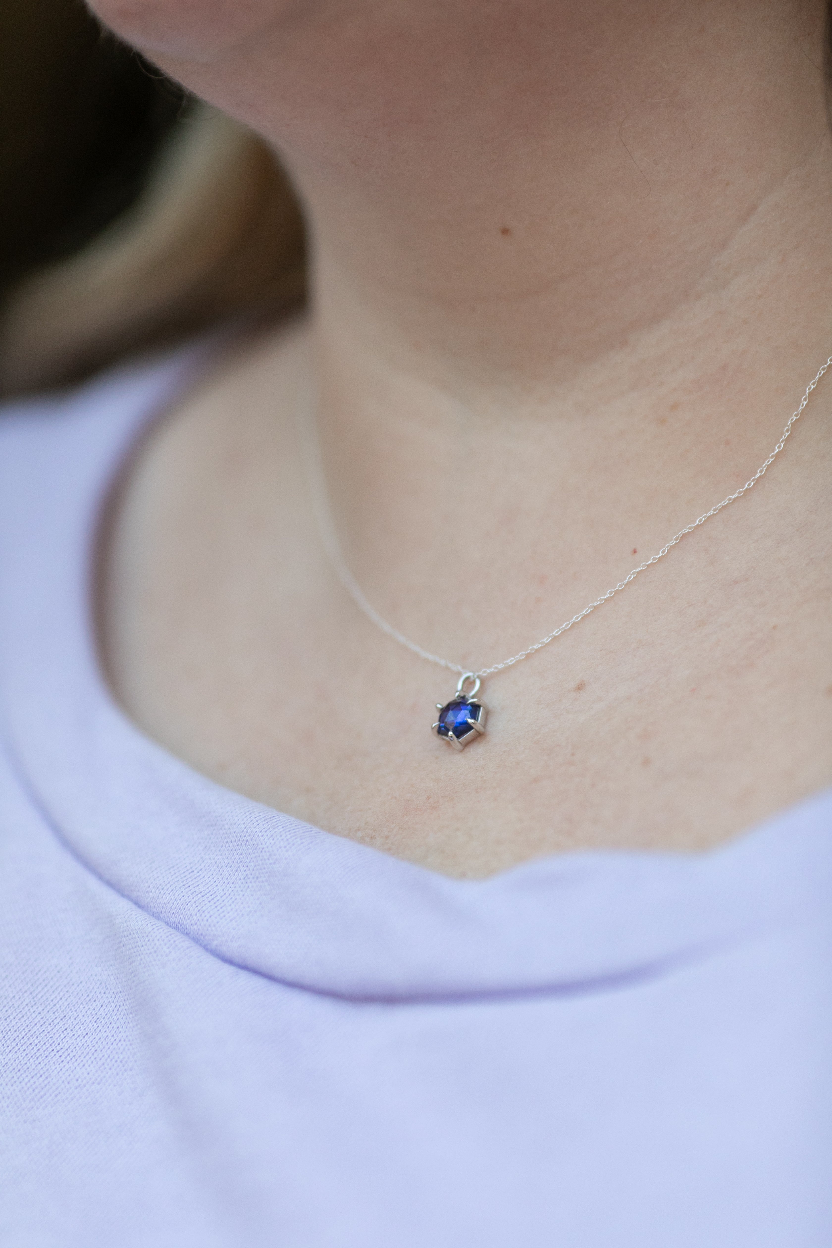 a hexagonal rose cut tanzanite set in a simple silver chain lays on a neck above a light lilac sweater