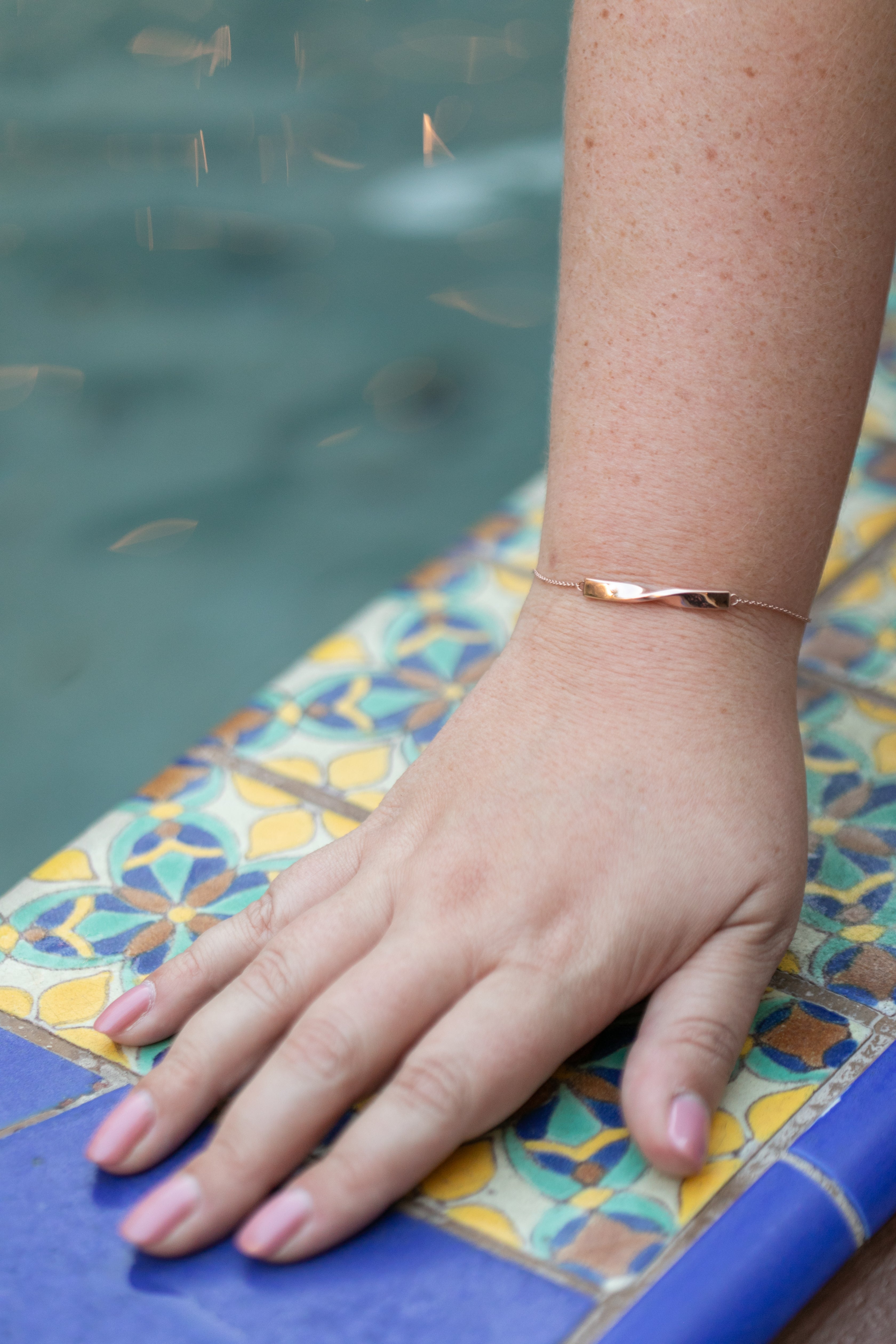 a plus size woman's hand rests on the side of a fountain over Spanish style tiles with a twist bracelet in rose gold on her wrist.