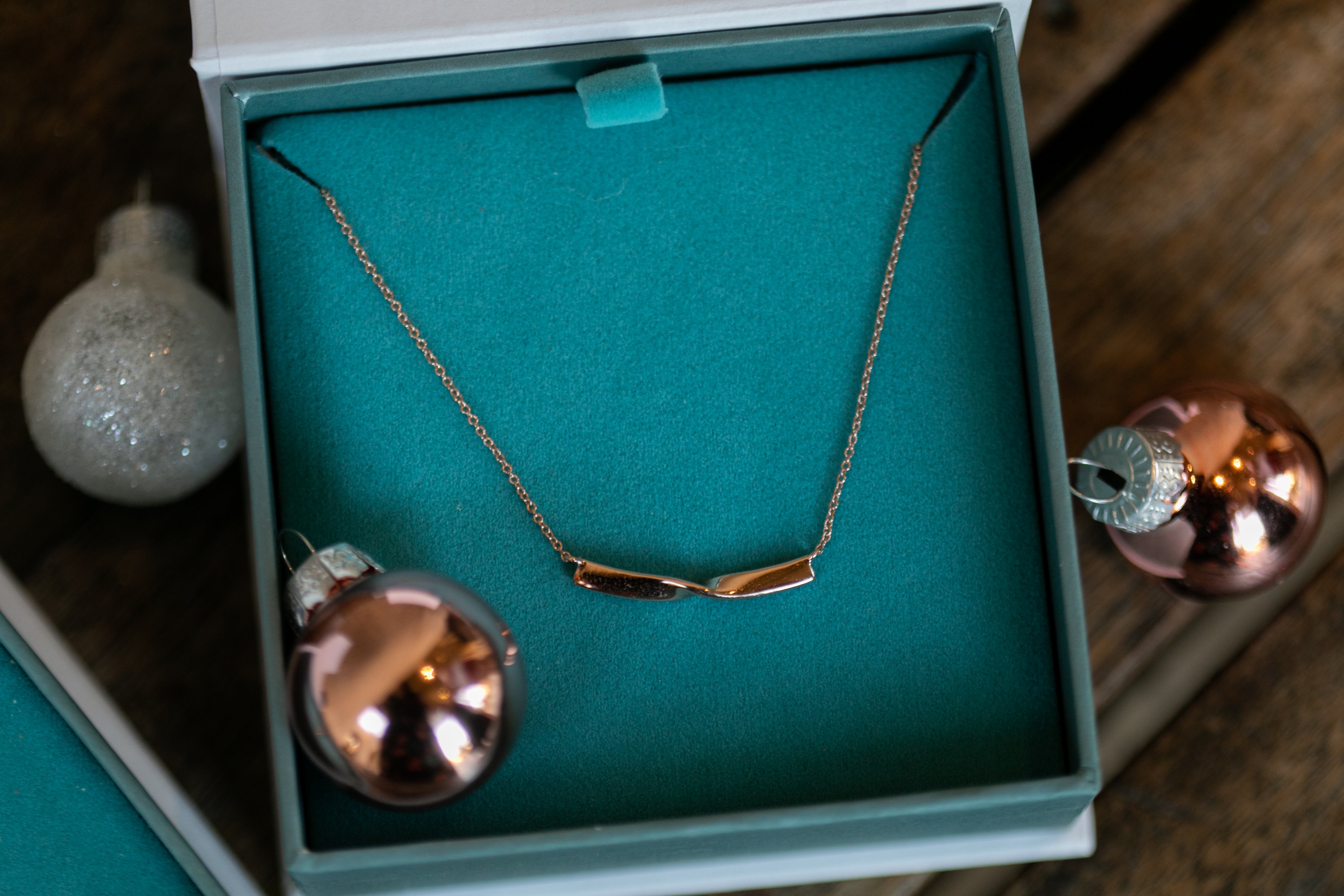 a close up of a rose gold twist necklace in a teal Chouette Designs branded jewelry box, surrounded by rose gold and glittery Christmas ornaments