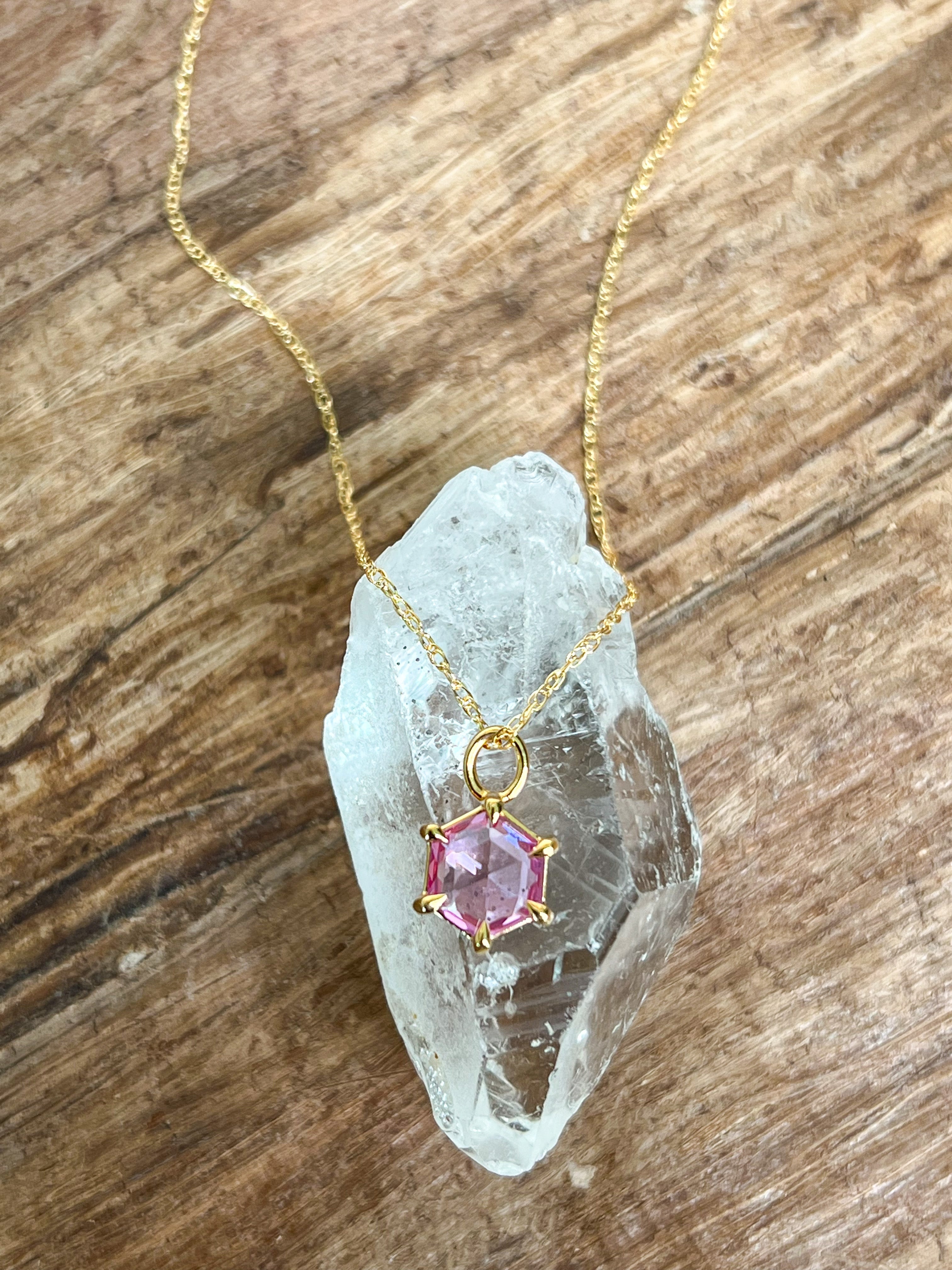 a close up shot of the gemstone pendant necklace in gold and pink spinel draped over a clear crystal. The spinel is a rose cut hexagonal shape on a delicate chain.
