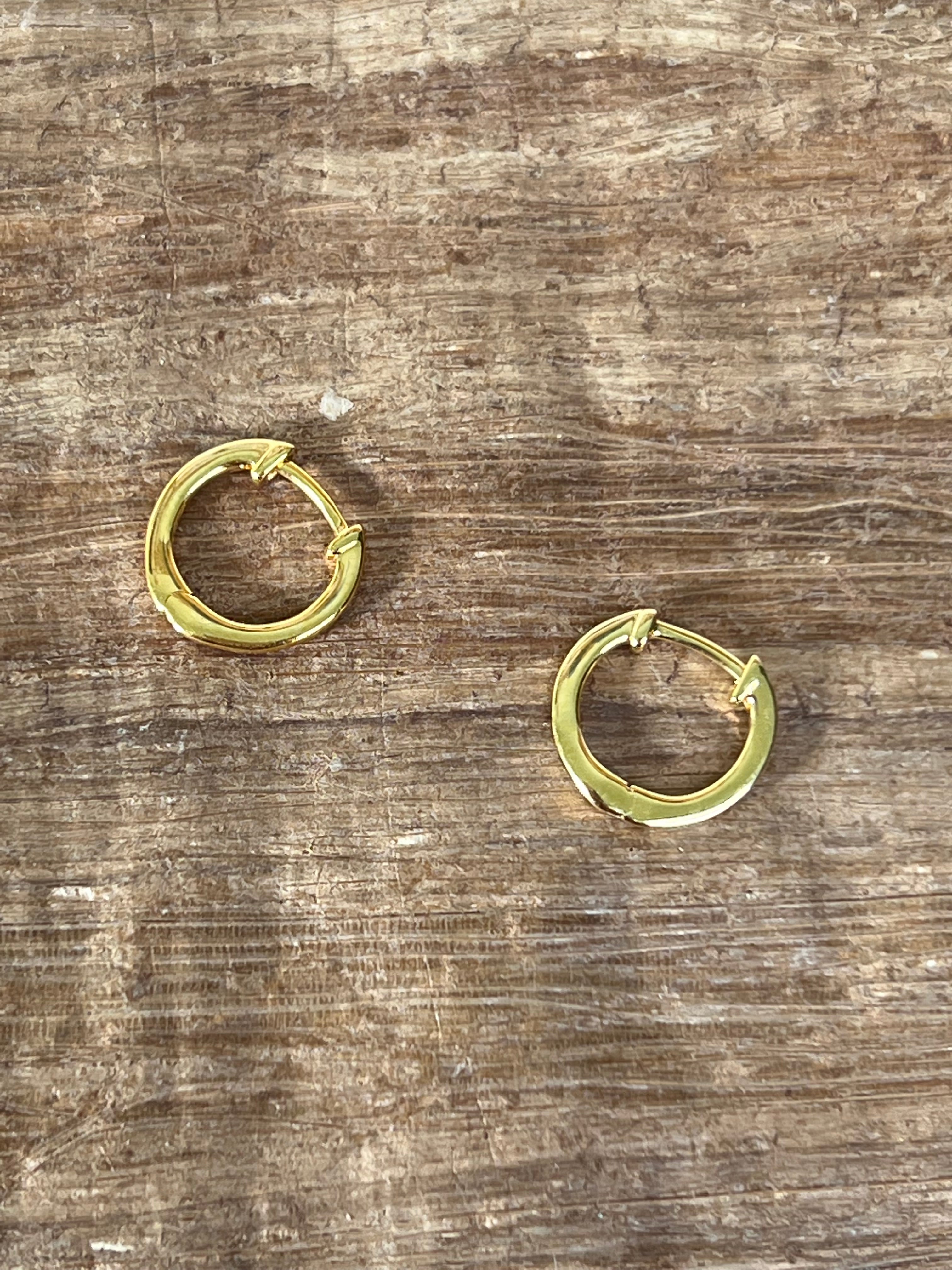 a detail image of two huggie hoop earrings in yellow gold laying on top of a wood grain backdrop.